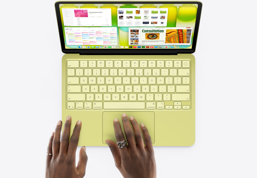 MacBook Neo citrusszínben, az érintőpadon egy felhasználó keze, a kijelzőn több különböző alkalmazás, köztük a Zene, a Naptár és a Microsoft PowerPoint ablaka.