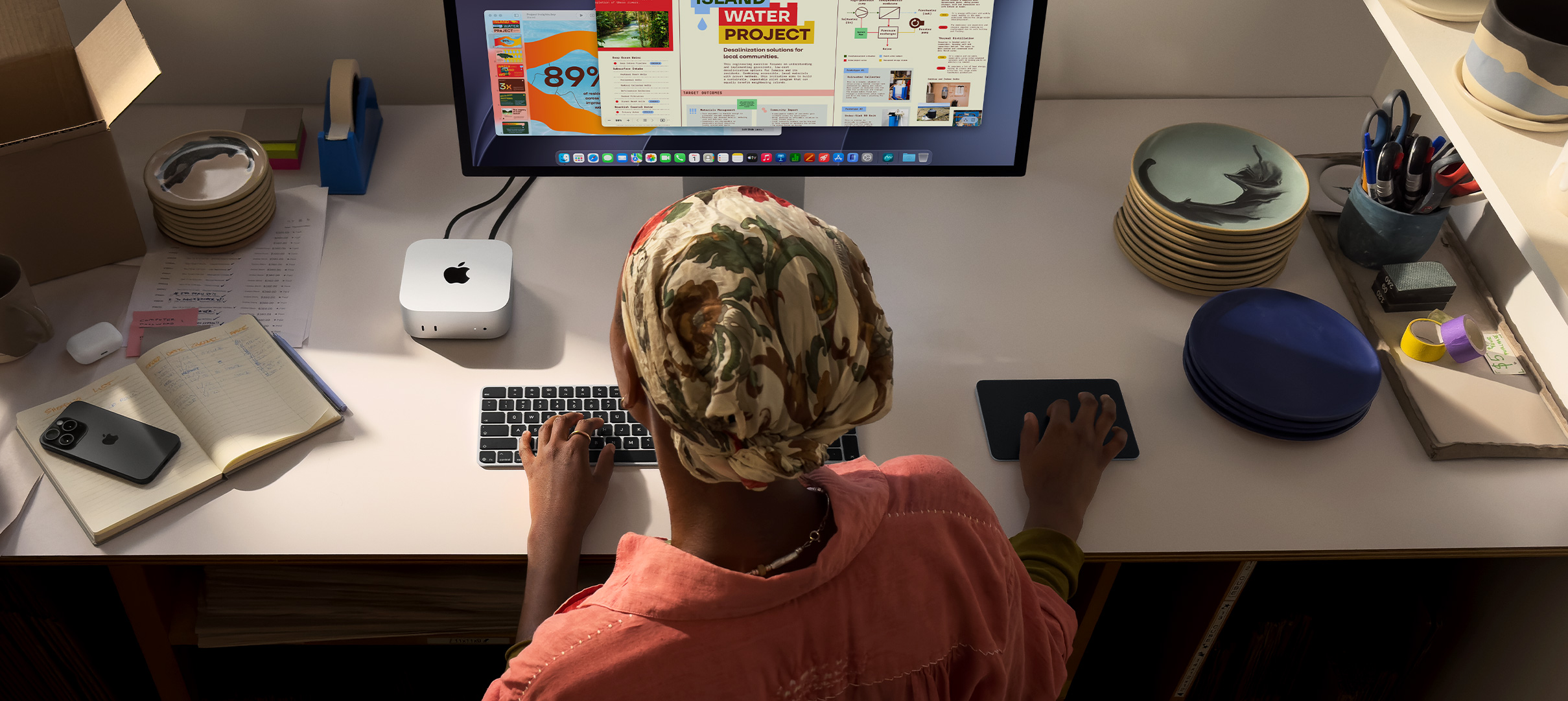Seseorang mengetik di meja dengan Mac mini, Studio Display terhubung, Magic Keyboard, Magic Trackpad, berbagai barang