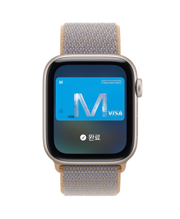 Apple Watch SE 3, 스타라이트 색상 알루미늄 케이스 및 ‘지갑’ 앱에 결제 카드 이미지가 표시되고 오른쪽 옆면 외관에 Digital Crown 다이얼이 보입니다. 샴브레이 블루(라이트 블루 그레이) 색상 스포츠 루프 밴드가 보입니다.