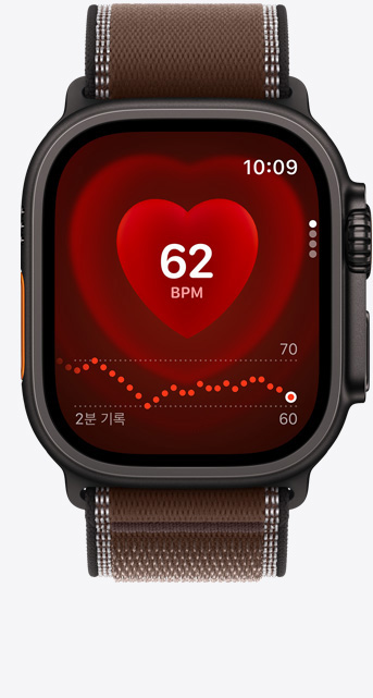 블랙 색상 티타늄 케이스의 Apple Watch Ultra 4. '심박수' 앱에 현재 분당 심박수가 표시되어 있으며 다크 엄버 색상(브라운) 트레일 루프 밴드를 장착