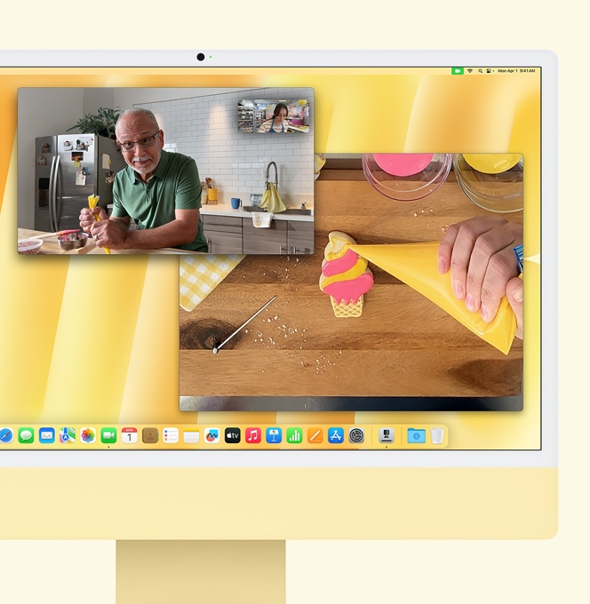 Žltý iMac. Ukážka Pohľadu na stôl. Obrazovka zobrazujúca cukrárku na videohovore s ďalším človekom. Jedna obrazovka zobrazuje cukrárku a tvár muža. Druhá obrazovka zobrazuje ruky zdobiace sušienku