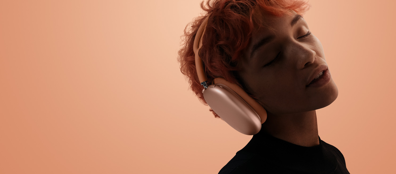 Persona ar pieliektu galvu un aizvērtām acīm ir uzlikusi AirPods Max 2 oranžā krāsā, uz oranža un rozā fona