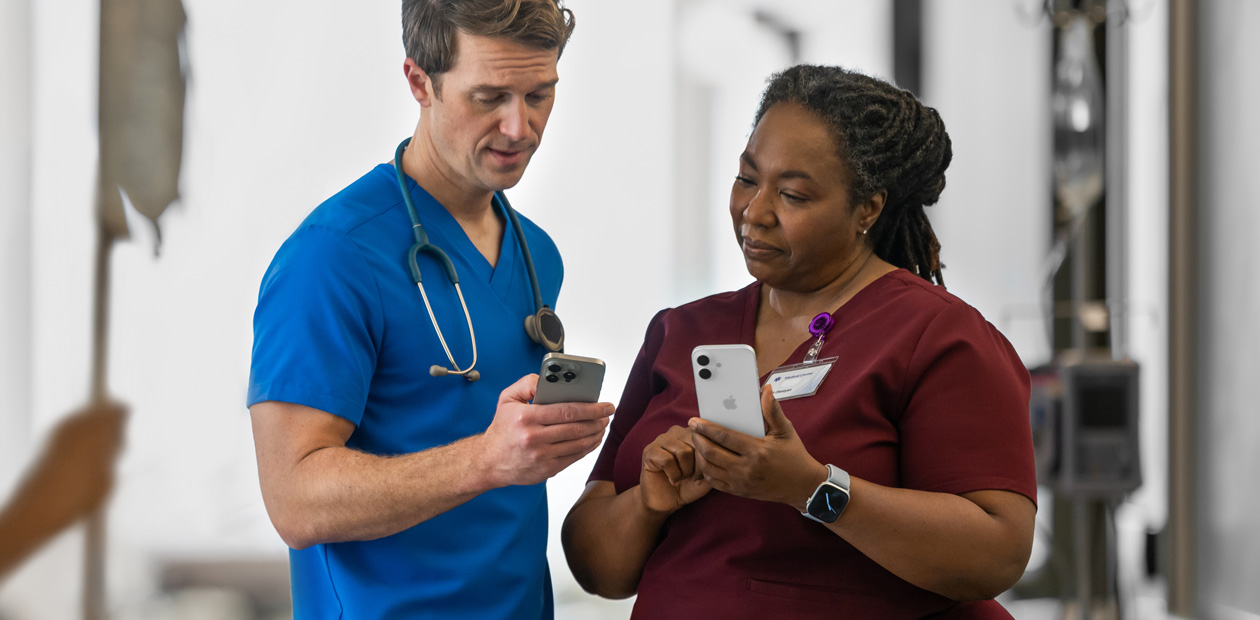 Un prestataire de soins en blouse bleue regarde son iPhone et montre quelque chose à sa consœur en blouse rouge, qui dispose, elle aussi, d’un iPhone.