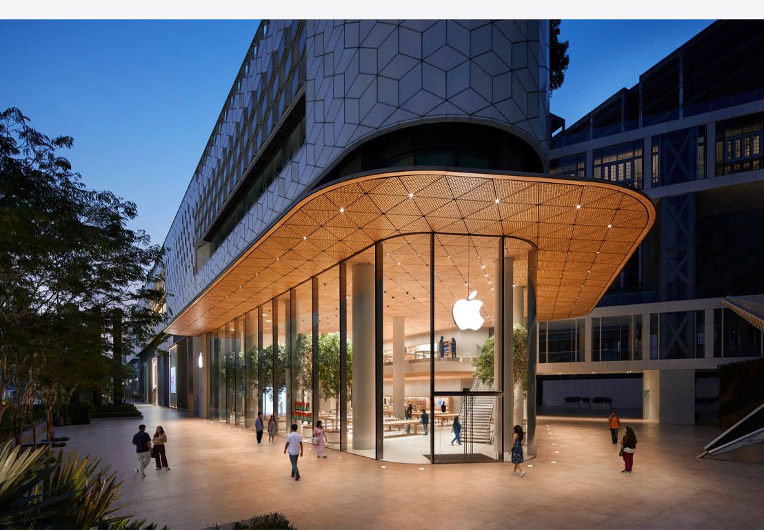 Apple Store de noche, ventanas de vidrio, interior de la tienda iluminada, logo de Apple centrado en la parte superior de la puerta de entrada.