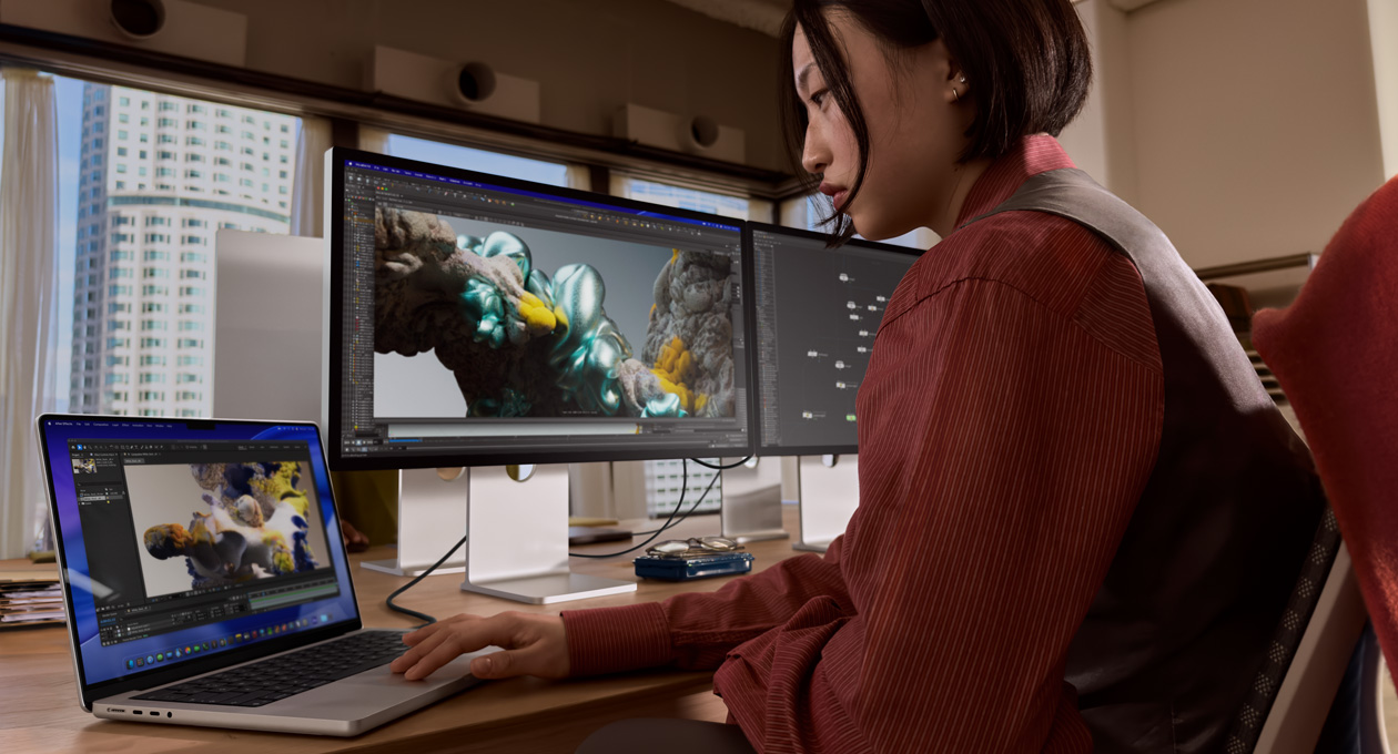 A person in an office setting works on a MacBook laptop that is connected to Studio Display XDR by a Thunderbolt 5 port